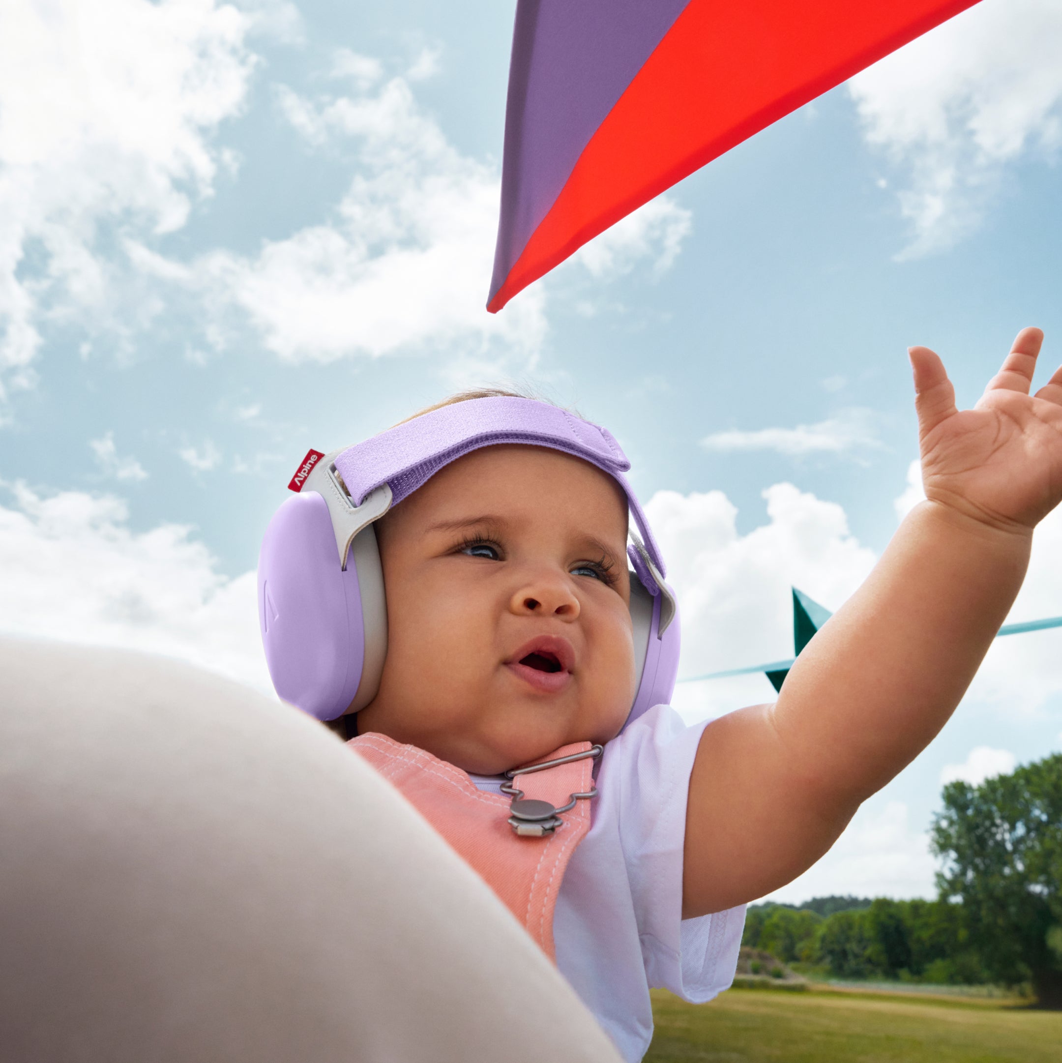 Baby wearing purple muffy baby earmuff and a pink shirt, looking up at a red kite against a blue sky.